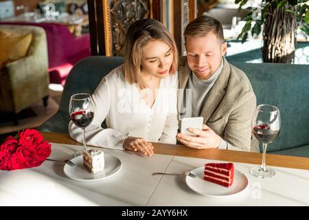Photo horizontale en grand angle de jeune homme et de jeune femme assis ensemble dans un café moderne en regardant quelque chose sur smartphone Banque D'Images