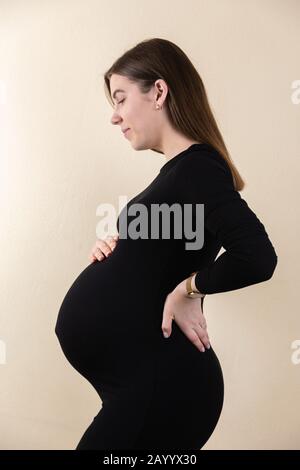 Vue latérale d'une jolie femme enceinte caressant son ventre en robe noire tout en se tenant sur fond vertical jaune Banque D'Images