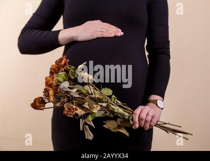 Vue latérale d'une jolie femme enceinte caressant son ventre et tenant un bouquet de roses sèches en robe noire tout en se tenant contre le backgrou jaune Banque D'Images