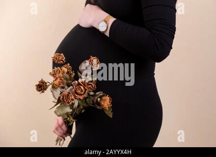 Vue latérale d'une jolie femme enceinte caressant son ventre et tenant un bouquet de roses sèches en robe noire tout en se tenant contre le backgrou jaune Banque D'Images