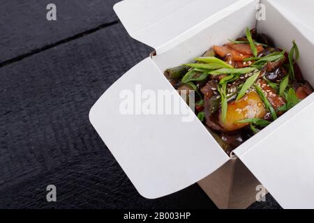 Nouilles avec porc et légumes dans la boîte à emporter sur table en bois, photo verticale Banque D'Images