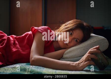 Jeune jolie femme vêtue de robe rouge posée sur le lit et regardant la vidéo sur smartphone le soir. Accueil soirée détente et repos concept Banque D'Images