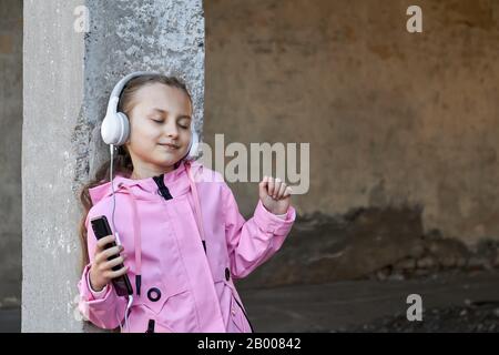 Une petite fille caucasienne dans un manteau rose écoute de la musique dans un casque contre un mur en béton. Fille dansant avec les yeux fermés. Casque sans fil Banque D'Images