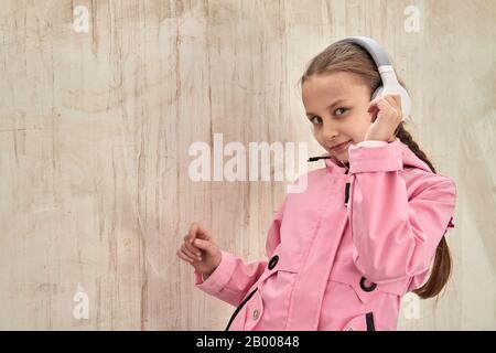 Une petite fille caucasienne dans un manteau rose écoute de la musique dans un casque contre un mur en béton. Fille dansant avec les yeux fermés. Profitez de la musique. Fil Banque D'Images