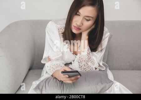 Une femme asiatique porte une robe de nuit blanche et un peignoir en satin à manches longues avec dentelle fleurie sur le canapé tard dans la nuit en utilisant un téléphone portable endormi et fatigué dedans Banque D'Images