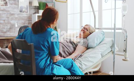 Dans la maison de retraite lumineuse, la vieille dame qui menait dans le lit d'hôpital parle avec une femme soignant. Banque D'Images