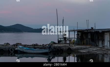 Zepko, Grèce - 26 avril 2019: Petit port au crépuscule avec petit bateau à ramer et cabine avec chaises et table et en arrière-plan quelques collines et rouge Banque D'Images