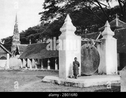 Thaïlande: Un moine novice se trouve à côté du grand gong de Wat Phra que Haripunchai, Lamphun, c.1920. Wat Phra que Haripunchai a été fondé en 1044 par le roi Athitayarat de Haripunchai sur le site du palais royal de la reine Chamathewi (Chama Thewi ou Chamadevi).La légende dit que les quartiers personnels de la reine sont enfermés dans le chedi de style Na LAN principal de 46 mètres de haut, recouvert de plaques de cuivre et recouvert d'un parapluie or ou d'une plée.Lamphun a été la capitale du petit Royaume mon de Haripunchai, mais culturellement riche, d'environ 750 ce à l'époque de sa conquête par le roi Mangrai en 1281. Banque D'Images