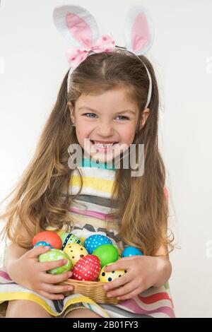 Jolie petite fille portant des oreilles de lapin et tenant des œufs de Pâques peints et souriant sur un fond blanc. Jour de Pâques. Un bébé heureux en vacances. Banque D'Images