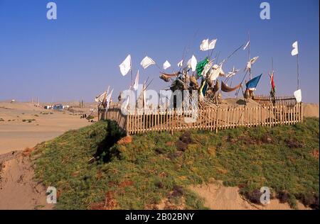 Chine : un tombeau dans les dunes de sable près de l'Imam Asim Mazar (sanctuaire en arrière-plan) dans le désert près de Khotan, province du Xinjiang. Le sanctuaire de l'Imam Asim et d'autres tombes se trouvent à 23 kilomètres de Khotan (Hotan) sur la route de la soie au sud. C'est le site d'un immense pèlerinage chaque mois de mai. L’Imam Asim a été l’un des premiers missionnaires islamiques à visiter cette région. Khotan retrace son histoire au moins aussi loin que le IIIe siècle av. J.-C., lorsque le fils aîné de l'empereur indien Asoka s'est installé ici. Banque D'Images