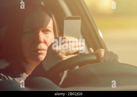 Femme d'affaires conduite de voiture et messagerie texte sur téléphone mobile, main tenant smartphone et volant de véhicule, mise au point sélective Banque D'Images