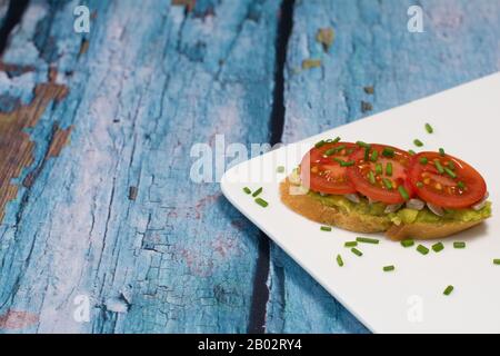 Un sandwich ouvert avec avocat étalé à l'ail, aux graines de tournesol, aux tranches de tomates cerises et aux ciboulettes en dés, sur une plaque blanche. Banque D'Images