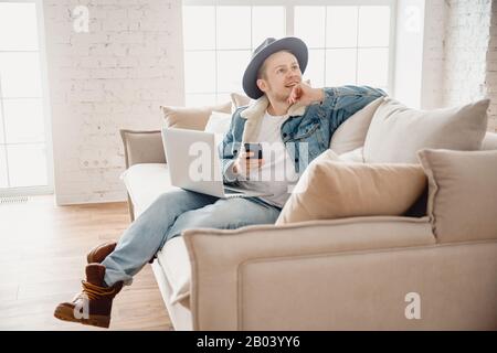 Homme barbu collègue hipster dans chapeau travail salle de séjour maison. Homme assis sur un canapé à l'aide d'un ordinateur portable et d'un téléphone portable. Fond de lumière flou Banque D'Images