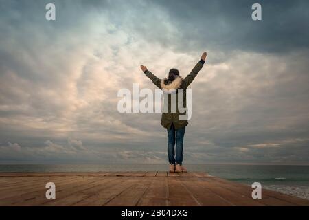 Femme portant un manteau parka debout par la mer avec ses bras levés, concept de style de vie. Banque D'Images