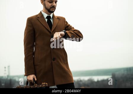 Portrait d'un jeune gars de trente ans, dans un style d'affaires, avec un sac en cuir dans ses mains, regardant sa montre. Contre le ciel, style de rue Banque D'Images