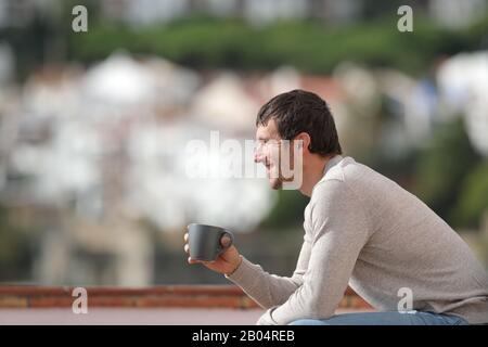 Vue latérale portrait d'un heureux homme tenant une tasse de café assis sur un banc en regardant loin dans une ville une journée ensoleillée Banque D'Images