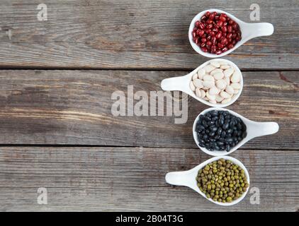 Ensemble de différentes variétés de haricots: Lima de bébé, petite noire, adzuki et mung dans des cuillères en céramique blanche (hors-d'œuvre) sur fond de bois, foc sélectif Banque D'Images