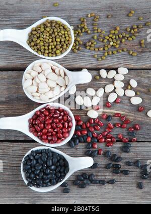 Ensemble de différentes variétés de haricots: Lima de bébé, petite noire, adzuki et mung dans des cuillères en céramique blanche (hors-d'œuvre) sur fond de bois, foc sélectif Banque D'Images
