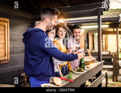 Les jeunes amis applaudissant et riant dans un bungalow en bois du village de camping - Les Personnes Heureuses ayant un dîner en plein air amusant barbecue et toaster le vin rouge - Banque D'Images