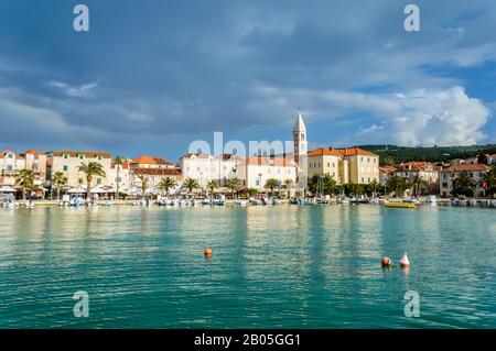 Supetar sur l'île de Brac près de Split, Croatie. Petite ville balnéaire avec promenade et port avec bateaux, palmiers, cafés, maisons et église. Touristes wa Banque D'Images