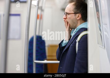 Homme fatigué dans les lunettes dans les transports publics. Jeune homme épuisé debout et bâillant dans le métro, endormi après le travail rentre à la maison, fond flou Banque D'Images