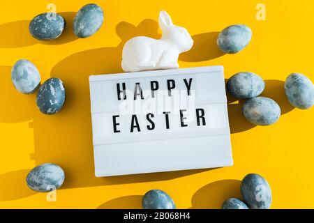 Œufs de Pâques peints et texturés bleus, lapin en plastique blanc et panneau décoratif lumineux avec les lettres Joyeuses Pâques sur un backgr de vacances jaune vif Banque D'Images