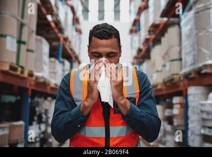 Jeune employé d'entrepôt africain malade soufflant le nez tout en travaillant avec un gilet de sécurité Banque D'Images