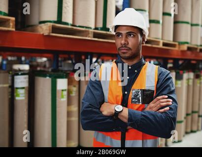 Portrait d'un jeune homme africain américain sérieux avec des mains repliées et un chapeau blanc debout dans l'entrepôt de fabrication tout en regardant loin Banque D'Images