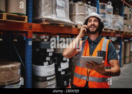 Beau homme heureux en casque blanc et gilet de sécurité tenant debout tablette numérique tout en parlant au téléphone regardant loin debout entre les marchandises dans Banque D'Images