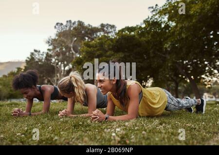 Jeune homme souriant trois femmes multiraciales diverses dans des vêtements de sport faisant des exercices de planche sur l'herbe verte dans le parc une journée ensoleillée Banque D'Images