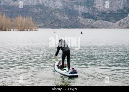 Un homme avec des vêtements chauds fait du paddle surf explorer dans un lac de montagne avec des bagages en haut de son conseil Banque D'Images