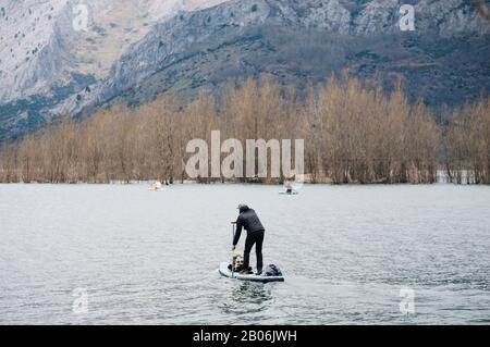 Un homme avec des vêtements chauds fait du paddle surf explorer dans un lac de montagne avec des bagages en haut de son conseil Banque D'Images