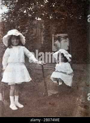 Archivistique, ancienne, noire et blanche photographie de deux jeunes filles dans un jardin, l'une d'elles assis dans un chariot à roulettes alors que la jeune fille tient la poignée du chariot comme si elle le tire. Les deux filles portent des robes blanches et des chapeaux blancs. Banque D'Images