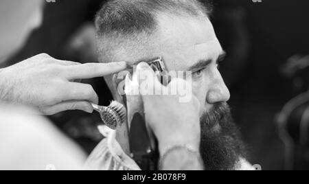 Vue latérale portrait d'un homme barbu adulte à poil sombre dans un barbier moderne pendant le processus de coupe des cheveux avec rasoir électrique. Outils et accessoires professionnels dans les mains de barber. Image en noir et blanc Banque D'Images