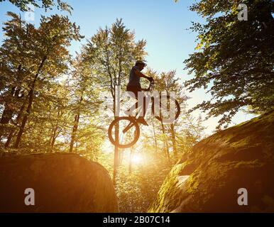 Jeune cycliste mâle sautant sur la bicyclette d'essai entre deux gros rochers, pilote professionnel faisant un tour acrobatique dans la forêt le jour ensoleillé. Concept de style de vie actif sport extrême Banque D'Images