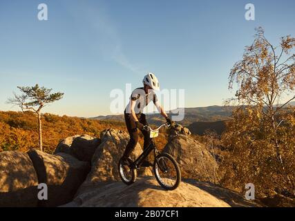 Les jeunes cyclistes masculins en équilibre sur le vélo d'essai haut de big boulder, rider stunt acrobatique faire sur soir d'été, ciel bleu et de la forêt sur l'arrière-plan. Concept de sport extrême de vie actif Banque D'Images