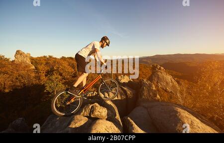 Cycliste professionnel en équilibre sur la bicyclette d'essai au-dessus de Big rock, cycliste mâle faisant acrobatique cascades le soir d'été, ciel bleu et forêt en arrière-plan. Concept de style de vie actif sport extrême Banque D'Images