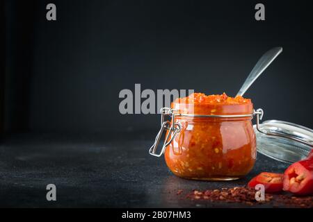 Sauce traditionnelle au piment chaud Jadjika, pâte harissa dans un pot en verre noir. Cuisine tunisienne, géorgienne et arabe. Orientation horizontale. Espace pour Banque D'Images