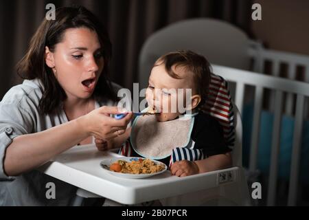 Mère nourrissant porridge à son bébé enfant garçon chambre intérieure pour enfants - Jeune maman en gris vêtements de maison et son fils manger assis dans le siège de bébé - Heureux Banque D'Images