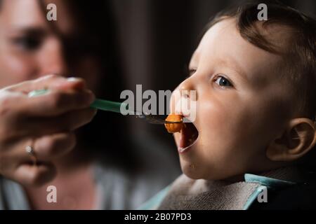 Mère nourrissant porridge à son bébé enfant garçon chambre intérieure pour enfants - Jeune maman en gris vêtements de maison et son fils manger assis dans le siège de bébé - Heureux Banque D'Images