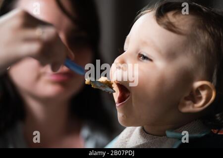 Mère nourrissant porridge à son bébé enfant garçon chambre intérieure pour enfants - Jeune maman en gris vêtements de maison et son fils manger assis dans le siège de bébé - Heureux Banque D'Images