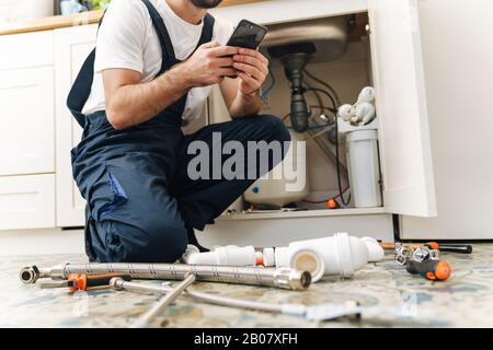 La photo courte du plombier de jeune homme travaille en uniforme à l'intérieur à l'aide d'un téléphone portable. Banque D'Images