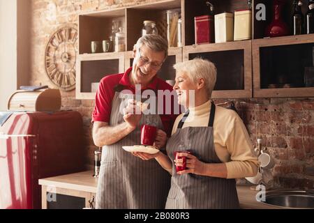 Portrait Du Couple Senior Boire Du Café Et Manger Des Biscuits Dans La Cuisine Banque D'Images