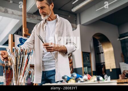 Artiste senior debout en studio picking paintbrush pour le dessin Banque D'Images