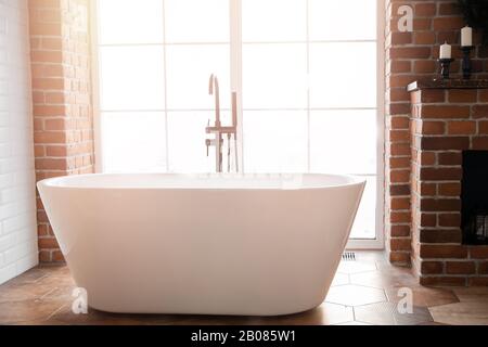 Salle de bains moderne avec baignoire en acrylique blanc sur pied et mixeur en mezzanine noir. Murs en briques, parquet en bois, lumière du soleil depuis la fenêtre Banque D'Images