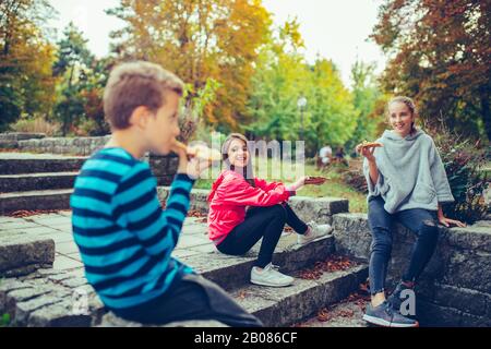 Trois enfants heureux de parler et de manger de la pizza à l'extérieur, s'amuser ensemble. Banque D'Images