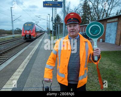 06 février 2020, Mecklembourg-Poméranie occidentale, Sülstorf: Le chauffeur de taxi de Berlin Michael Böhmke est debout sur la plate-forme devant la gare "HIS" de Sülstorf, portant un chapeau de chemin de fer et une truelle. En arrière-plan, le express régional des arrêts de la Deutsche Bahn. Il y a deux ans, Böhmke a acheté la gare sur la ligne de chemin de fer de Ludwigslust à Schwerin lors d'une vente aux enchères sur Ebay. Peu à peu, il veut maintenant étendre et rénover le bâtiment; il a déjà recueilli de nombreux éléments de collection relatifs à l'histoire ferroviaire. Photo: Jens Büttner/dpa-Zentralbild/ZB Banque D'Images