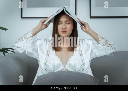 Une femme asiatique porte une robe de nuit blanche et une robe de satin à manches longues avec dentelle fleurie souffrant de fatigue oculaire livre de lecture assis sur un canapé dans la nuit à la maison. Banque D'Images