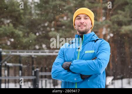 Portrait d'un jeune homme sportif souriant et confiant dans un chapeau traversant les bras contre un terrain de sport en plein air en hiver Banque D'Images
