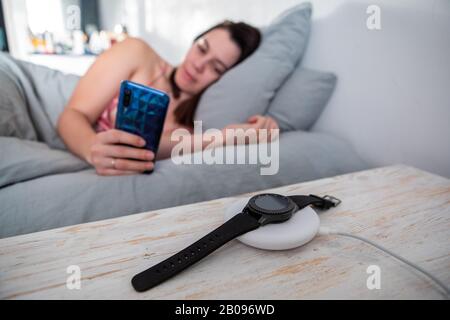 femme surfant sur internet sur le téléphone portable posé dans le lit smart watch sur le chargeur sans fil Banque D'Images